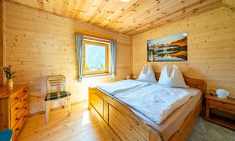 A cozy wooden bedroom with a large bed and a window that lets in daylight. A landscape painting hangs on the wall, and there is an armchair as well as a dresser in the room.