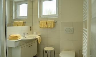 A modern bathroom with bright tiles and large windows. There are yellow towels on the washbasin and a small stool stands next to it.