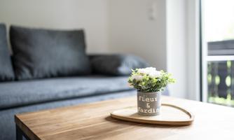 A cozy living room with a gray sofa and a wooden table. On the table sits a small plant pot with the inscription "Fleurs & Jardin".