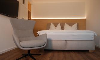 A modern room with a comfortable bed and a cozy armchair. The soft colors and warm lighting create an inviting atmosphere.