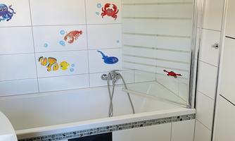 A modern bathroom with a bathtub and a shower. Colorful fish and crabs decorate the white tiles.