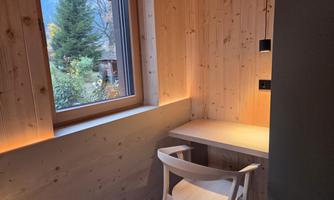 A bright room with wooden walls and a window. A chair is at the table, and light accentuates the walls.