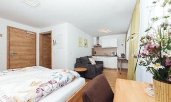 A modern room with a bed, a seating area, and a small kitchenette. The walls are light and decorated with plants.