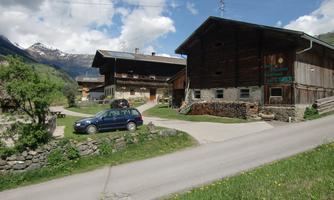 A traditional country house with a wooden facade in a rural setting. In the foreground, there is a road and a blue car, surrounded by green landscape.