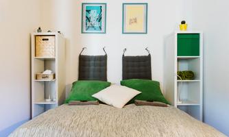 A modern bedroom with a double bed and green cushions. There are pictures hanging on the wall and shelves with decorative elements.