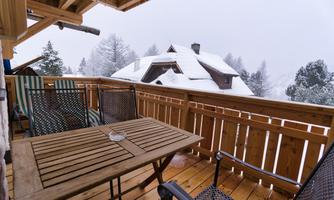 A cozy balcony with wooden furniture and a view of snow-covered trees. The surroundings appear calm and wintry.