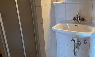 A simple bathroom with a shower, a sink, and a mirror. The walls are tiled with white tiles.