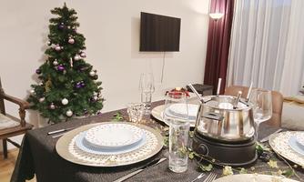 A festively set table with dishes, glasses, and a fondue set. In the background stands a decorated Christmas tree.