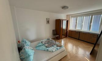 A simple bedroom with a white bed and colorful pillows. Large windows provide plenty of daylight, and wooden wardrobes complement the decor.