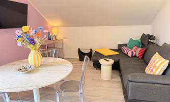 A stylish living room with a gray sofa and colorful cushions. On the table is a vase with flowers and the room has a bright, inviting atmosphere.
