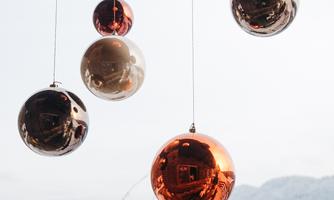 Hanging, shiny Christmas ornaments in various colors against a snowy landscape. In the background, houses and snow-covered mountains can be seen.