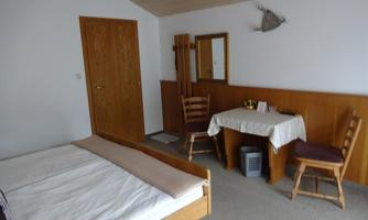 A simple room with a double bed, a table, and two chairs. The walls are light, and there is a mirror as well as a wooden decoration.