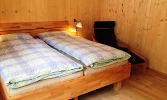 A cozy bedroom with a wooden double bed and blue bedding. Next to the bed is a black chair with a small lamp.
