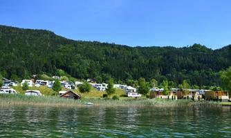 A peaceful lake with green shore areas and campsites in the background. The surroundings are surrounded by wooded hills and the sky is clear and blue.