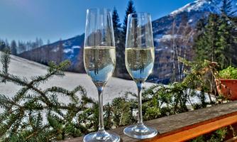 Two glasses of sparkling wine are placed on a wooden board, surrounded by pine branches. In the background, snowy mountains and a blue sky are visible.