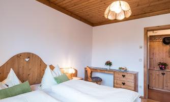 A cozy bedroom with two single beds and wooden decor. The room is bright and inviting with a small desk corner.