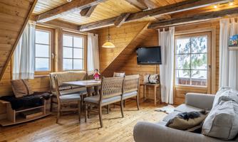 A cozy living space in a wooden house with a dining table, chairs, and a sofa. Large windows let in plenty of light and offer a view of nature.