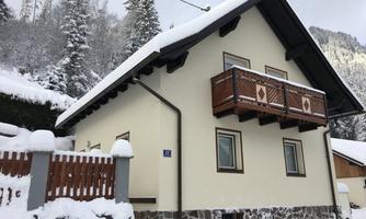 A cozy house in the snow with a balcony and a wooden veranda. The surroundings are enveloped by snow-covered trees.