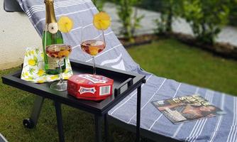 A cozy outdoor area with a lounge chair. On a table are two cocktails, a bottle of champagne, and a magazine.
