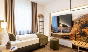 A modern hotel room with a large window and bright curtains. There is a comfortable bed, a television, and a decorative wall featuring a landscape motif.