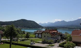 A picturesque landscape with a clear lake and gentle mountains in the background. In the foreground, there are green meadows and a small residential area with colorful houses.