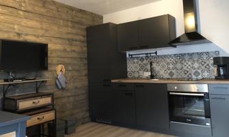 A modern kitchen with dark cabinets and an elegant backsplash. The space is minimally and brightly furnished, with a television and wooden details.