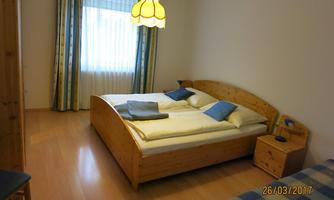 A simple bedroom with a double bed and blue pillows. The room has large windows with curtains and a cozy atmosphere.