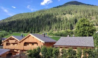 A picturesque mountain landscape with wooden chalets in the foreground. Green forests and a blue sky give the scene an idyllic atmosphere.