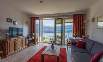 A cozy living room with a large window front and a view of a lake. The room is furnished in warm colors and features modern furniture.