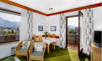 A bright kitchen-living area with a table, chairs, and a cozy seating area. The view shows mountains and a green landscape through the window.