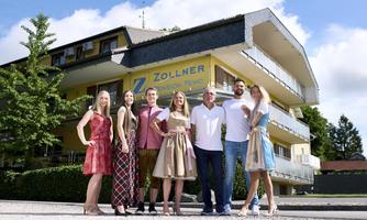 A group of seven people stands in front of a building named "Zollner". Trees and a blue sky can be seen in the background.