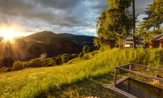 A picturesque view of a green meadow with mountains in the background. The sun rises behind the mountains and casts warm light on the landscape.