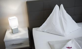 A clean bedroom area with a cozy bed and artfully folded towels. Next to the bed is a modern lamp on a nightstand.