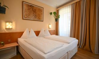 A cozy bedroom with a double bed and white bedcovers. The windows are fitted with curtains, and there are plants that create a pleasant atmosphere.