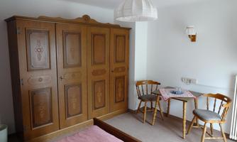 A simple room with a wooden wardrobe and a small table with two chairs. The floor is carpeted and the bed has a pink duvet.