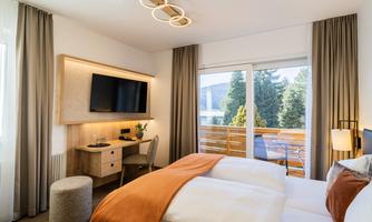 A modern hotel room with a double bed and a desk. Large windows offer a view of the green surroundings.