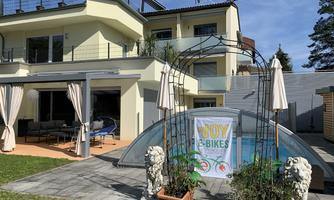 A modern house with a pool and a cozy outdoor area. Adjacent to it are two plants and a banner for e-bikes.