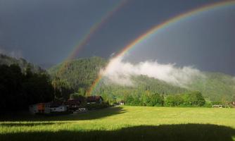 An impressive rainbow stretches over a green landscape with gentle hills. In the background, there are dark clouds and a clear view of nature.