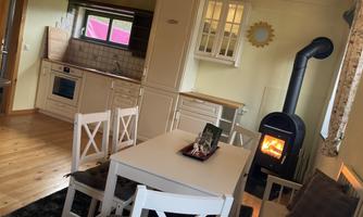 A cozy kitchen with a table and white chairs. A wood stove radiates warmth, while large windows let in natural light.