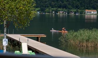 A calm lake with a wooden pier and green shore plants. Two people are paddling in a boat, while some houses can be seen in the background.