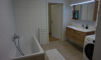 A modern bathroom with a bathtub, a sink, and a washing machine. The walls are tiled with white tiles and the lighting is bright and inviting.