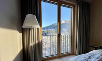 A bright room with a large window that offers a view of snow-covered mountains. The window is dressed with curtains, and a floor lamp and a bed are positioned in the foreground.