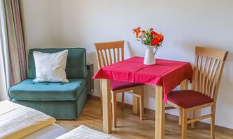 A cozy living area with a green armchair and a small wooden table. On the table is a vase with colorful flowers.