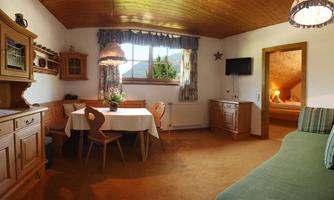 A cozy living space with wooden furniture and a round table. The windows let in a lot of light and offer a view of nature.