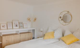 A bright bedroom with a double bed, white bed linens, and yellow pillows. A round mirror hangs on the wall, and decorative pictures are placed on a shelf.