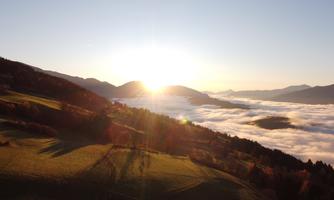 A stunning landscape with gentle hills and fog covering the valleys. The sun rises behind the mountains, creating a beautiful light ambiance.