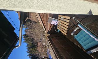 A small balcony with a table and chairs. In the background, there are trees and a blue sky.