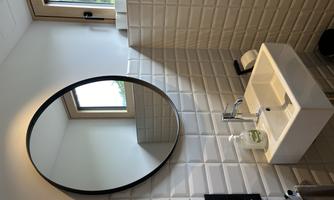 A modern bathroom with a white sink and a round mirror. The walls are tiled with bright, square tiles and there is a window that lets in daylight.