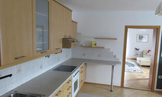 A modern kitchen with light wooden furniture and a window. In the background, the living room is visible.