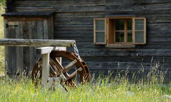 A historic wooden house with a window and a water feature. In the foreground is a water wheel, surrounded by green grass.